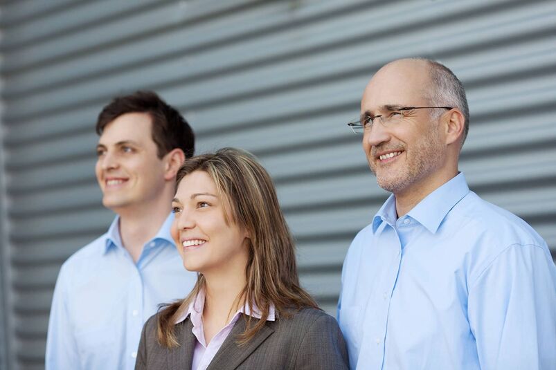 Zwei Generationen - ein Team - Von links: Ein junger Mann im hellblauen Hemd, eine junge Frau im Blazer, ein etwas älterer Mann im hellblauen Hemd schauen mit freundlichen Blick in eine Richtung.